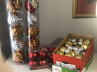 Photo showing two long tubes containing 12 large metallic gold and silver ball ornaments each, a box of 4 medium red matte ball ornaments, and an open box of assorted gold and silver plastic ornaments with different finishes.