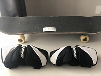 Photo showing the black grip tape side of the skateboard with wheels visible, accompanied by the pair of black and white Brine elbow pads.