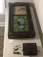 Front view of dark wooden antique wall clock with painted glass panel showing waterfall scene and Roman numeral dial, with separated mechanical parts beside it.