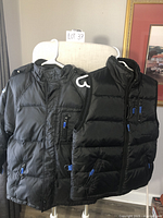 Two black youth GAP outerwear items on hangers, a down-filled jacket with hood and a matching vest, both with blue zipper pulls.