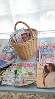 Wicker basket with magazines inside and laid out around it on a table. Magazines include Vogue, French Country, Cook's, Better Homes and Gardens, and various knitting and decorating magazines.