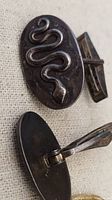 Close-up of one cufflink front showing raised snake design and part of toggle back with patina and tarnish visible on silver surface.