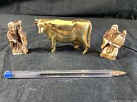 Front view of two bronze-tone figurines and one golden cow figurine placed on black cloth with pen for scale.