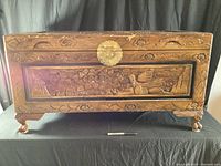 Front view of camphor wood trunk displaying large carved floral panel, bird motif, brass lock, and carved feet.