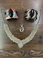 Top-down photo showing two distinct cuff bracelets with large black oval stones, a clear oval pendant with gold-tone metal wrapping, and a wide silver-tone rhinestone necklace