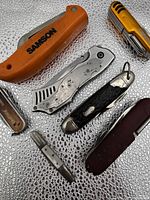 Top view of 7 assorted vintage cutting tools arranged compactly showing different sizes, colors and styles of pocket knives and multi-tools on a water-drop textured surface.