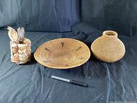 Three vintage First Nations baskets displayed on black fabric background, including flat woven tray, round bulbous basket, and small bark strip basket with fiber ties. Pen included for scale.
