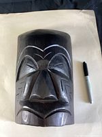 Front photo of the dark stained wooden Mosquito Mask with carved facial features and mosquito design elements next to a Sharpie for size reference.