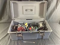 Top view of vintage fishing reels inside a grey plastic tackle box with the lid open. Showcases the collection and condition of reels.