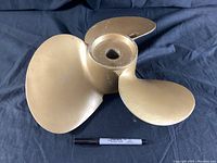Front angled view of the golden coloured three-blade boat propeller with a pen for size reference.