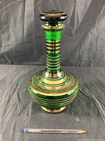 Front view of green Bohemian glass decanter with gold and red stripes and stopper on black background.