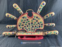Front view of the Indian carving set showing six knives arranged in a semi-circle on a red wooden stand with gold, green and blue floral enamel decoration.