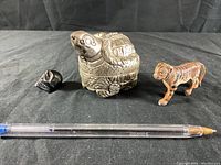 All three metal figurines displayed on black cloth with a pen for scale: skull, silver turtle, and big cat