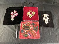 Four embroidered fabric panels laid out on a black surface, three with floral designs on black and red velvet and one with a bird motif on red cloth.