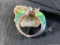 Front view of a silver-toned bracelet with two parrots facing each other, showing green enamel details on their wings and textured metal work on the heads and body, on a black background.
