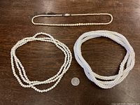 Four vintage pearl necklaces displayed on a wooden surface with a quarter for size reference. Three necklaces in a group: one long single strand with silver clasp, one triple strand creamy pearls, one triple strand white pearls.