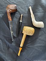 Four used pipes including dark wood, white meerschaum or clay, corn cob, and a black stem piece photographed on black fabric.