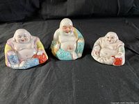 Front view of three seated porcelain Buddha figures showing distinct colorful robes and joyful expressions