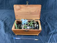 Open wooden jewelry box filled with assorted jewelry, placed on a black cloth with a pen for scale.
