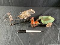View of both silver man cart and toy rooster cart with black background and pen for scale