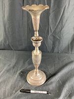 Full view of the tall silver-tone candle holder with scalloped trumpet-shaped top, mid cup, and circular base against a black fabric backdrop with a Sharpie pen for scale.