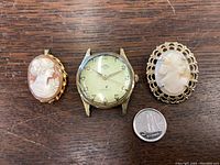 Photo showing two cameo brooches and one Solar automatic watch with quarter for scale on wooden surface.
