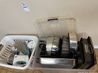 One plastic storage tote filled with metal baking pans, mixing bowls, cookie sheets, and wire racks, along with a white laundry basket containing miscellaneous baking utensils and accessories.