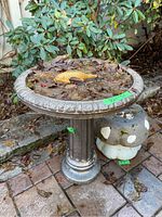 Concrete bird bath filled with leaves, featuring a yellow fish ornament and red berries, placed on a patio with bushes in the background.