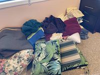 Multiple folded towels in various colors and fabric cushions with tropical leaf and striped designs piled on floor