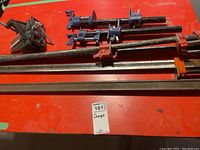 Photo of heavy metal clamps and a metal vise on a red surface. Several bar clamps in orange and red, two blue pipe clamps, and a black metal vise are visible.