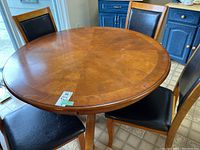 Round wood dining table with visible scratches and wear on the surface, surrounded by four matching chairs with dark seat and back upholstery.