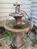 Concrete two tier outdoor fountain is shown in full view with power cable lying on the top basin and fountain center.