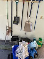 Various garden tools including shovels, broom, and rack hung on wall with items stored below including containers and bins.