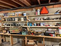 Wide view of basement shelves filled with assorted hardware, plastering tools, nails, fittings, and lighting equipment including a red emergency reflector triangle.