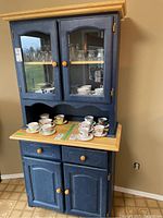 Front angled view of blue and natural wood hutch showing glass doors with china inside and storage cabinets below.