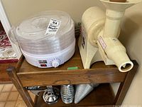 Photo showing Salton food dehydrator with multiple clear stackable trays and Champion juicer side by side on wooden cart, metal grinder visible below