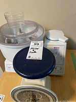 Front view of the vintage Way Rite kitchen scale with blue platform placed in front of Bosch Universal mixer. Mixer bowl and lid on top. Tag showing lot number 99 and text: "Mixer scale".