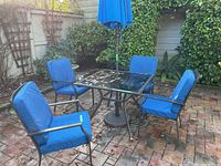 Full patio set including table, four chairs with blue cushions, umbrella, and base pictured outdoors on a brick patio surrounded by plants and fences.
