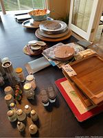 Photo showing stack of wooden trays, chargers, salad plates, melamine bowls, various salt and pepper shakers, and decorative trays on a table.