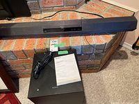 Sony black soundbar positioned on a brick surface with power cord visible. Accompanied by black rectangular subwoofer and instruction manual on top.