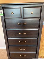 Front view of tall boy dresser showing 7 wooden drawers with 2 smaller top drawers and 5 larger drawers below, all with gold-colored hardware.
