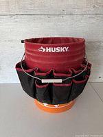 Front view of the Husky bucket organizer showing red upper fabric with logo and multiple black pockets on the perimeter.