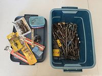 Wide top-down view showing storage tote with various drill bits including large yellow spade bits, hole saw, and many metal auger bits inside the tote and on side tray.