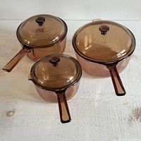 Set of three amber VISION Corning Ware glass pots with handles and lids displayed on a white wooden surface