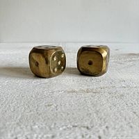 Front and side view of two brass dice showing standard dot markings, edges rounded, on flat surface.