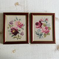 Two framed embroidery artworks side by side showing pink and purple floral designs on beige fabric.