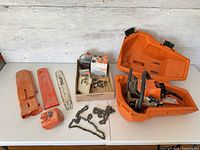 Full overview of disassembled chainsaw parts laid on white surface including case, bars and chains, and box of small parts.