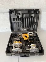 Open black hard plastic carrying case containing three router bases, two silver DeWalt bases and one yellow base with attached clear plastic guard and handles, all resting in custom foam compartments.