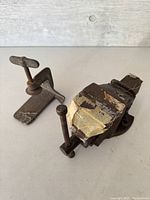 Two vintage metal clamps on a light surface, showing a small workbench vise and a C-clamp style clamp with signs of wear and tape residue.