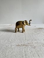 Front and side views of the vintage brass elephant figurine with trunk up on a textured neutral surface under natural light.